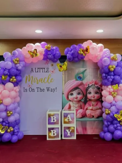 A sweet Krishna-themed baby shower decoration with the message "A Little Miracle Is On The Way." The setup includes a balloon arch in purple and pink, a themed backdrop, and "BABY" blocks.
