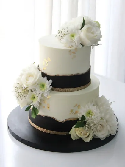 A timeless black, white, and gold engagement cake. This two-tiered design features a torn-edge effect and is adorned with fresh white florals for a modern and sophisticated look.