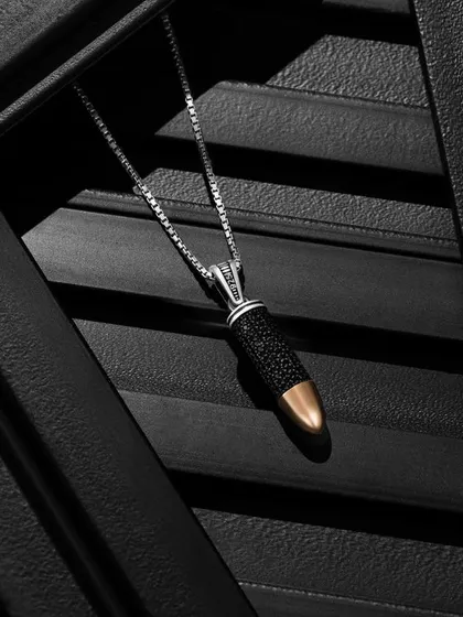A close-up of a bullet-shaped necklace, captured on dark, textured panels. The lighting is focused to highlight the intricate details and contrasting materials of the piece, creating a moody and masculine aesthetic.