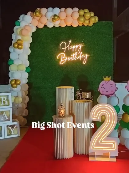 This rooftop birthday setup for a second birthday includes a balloon arch, a green turf backdrop with a neon sign, 'BABY' blocks, and a large light-up number '2'.