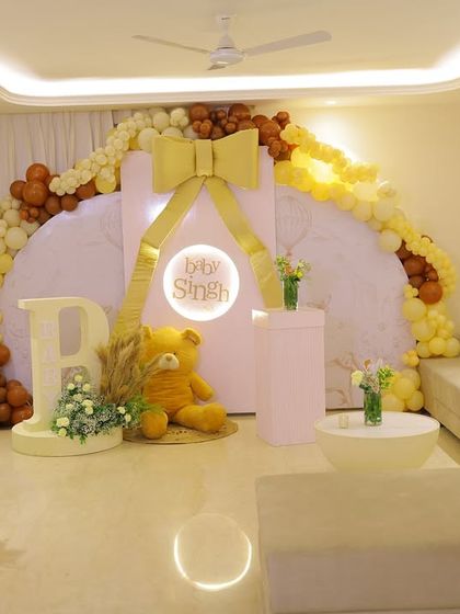 A wide view of an elegant baby shower setup in a living room. The decor features a large circular backdrop with a golden bow, a beautiful balloon arch in neutral tones, and a light-up sign for "Baby Singh."