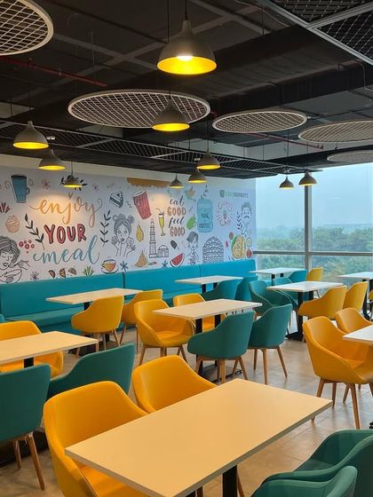 The vibrant cafeteria at the Cube Highways office, designed to be a refreshing breakout space. Colorful chairs, fun wall graphics, and modern lighting create an energetic and cheerful atmosphere.