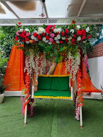 A beautifully decorated swing (jhoola) for a mehndi or haldi function. The swing is adorned with red, pink, and white flowers, with an orange drape in the background.