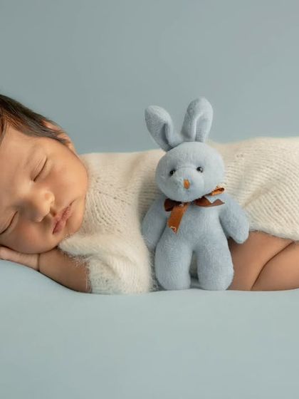 A newborn in a white bunny outfit with a fluffy tail sleeps on a light blue background next to a small blue stuffed rabbit.