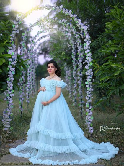 A dreamy solo portrait under a floral archway. The mother-to-be is wearing a light blue ruffled gown.