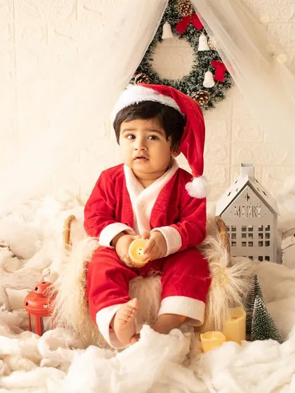 A baby Santa sits in a snowy, festive setup complete with a miniature house and reindeer.