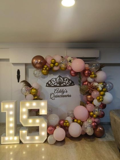 A Quinceañera (15th birthday) setup for a home party, with a pink and rose gold balloon circle, a custom backdrop, and light-up numbers.