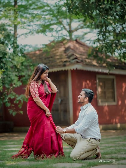 A classic romantic gesture during a pre-wedding shoot at a temple. The groom-to-be kneels before his partner, who is dressed in a beautiful red saree, creating a timeless and chivalrous photo.