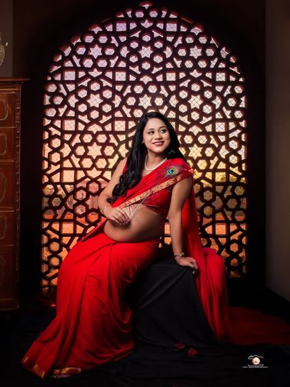 A joyful portrait of the expecting mother in a red saree, looking up with a smile. The warm light from the patterned window screen illuminates her, highlighting her happy expression and the traditional details of her look.