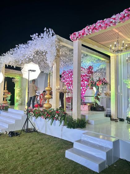 A beautiful stage setup for an outdoor wedding, featuring a canopy of white and pink florals. The design is being prepped by the team, showing the care that goes into every detail.