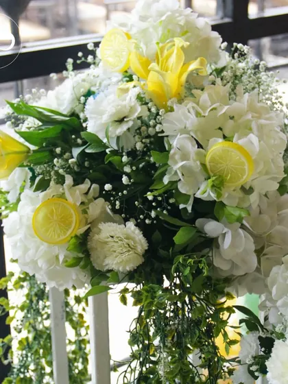 A beautiful floral arrangement featuring white hydrangeas, yellow lilies, and lemon slices. The cascading greenery adds a touch of drama and elegance to the citrus theme.