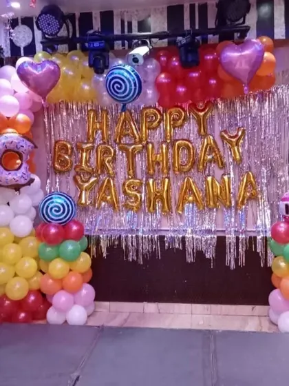 A vibrant candy-themed birthday decoration. This setup includes a silver tinsel backdrop, 'Happy Birthday' foil letters, and balloon pillars topped with lollipop and donut foil balloons.