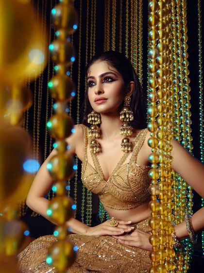 A striking shot of Manasa Varanasi for GNG Magazine, styled in a golden lehenga with dramatic, oversized jhumkas. The composition uses a beaded curtain to frame the subject, creating a rich, textured, and opulent portrait.