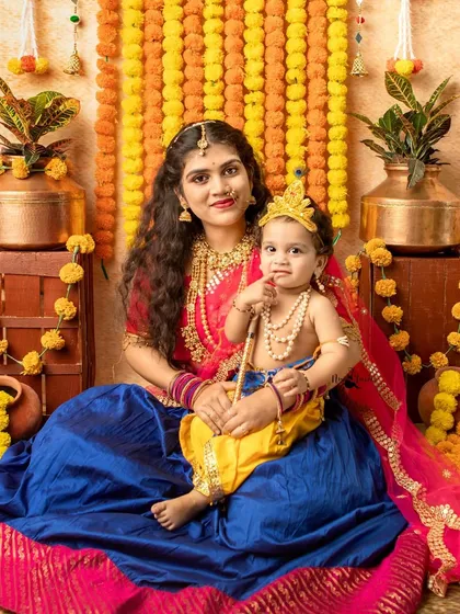 A beautiful portrait of a mother dressed as Yashoda with her little Kanha, set against a backdrop of vibrant marigold garlands.