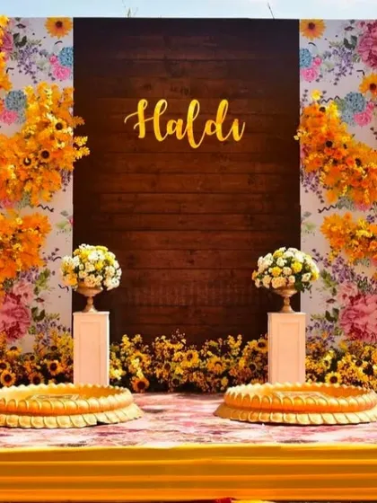 A close-up of a Haldi backdrop featuring a wooden panel with 'Haldi' written on it, framed by a beautiful arrangement of sunflowers and printed floral wallpaper.