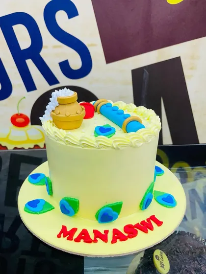 A custom cake celebrating Janmashtami, themed around Lord Krishna. This design includes a fondant flute, a pot of butter, and peacock feathers.