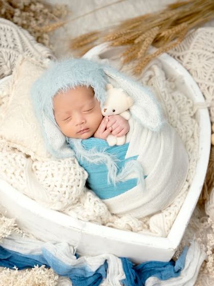 A wide shot showing a complete newborn setup. The baby is sleeping peacefully in a heart-shaped basket, surrounded by soft fabrics, wooden beads, and dried botanicals for a rustic and gentle feel.