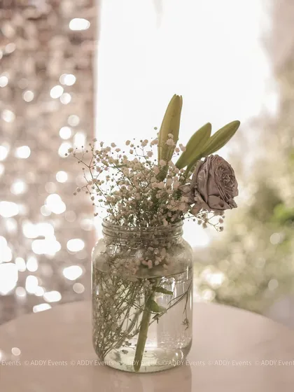 A simple yet beautiful decor detail. A silver rose and baby's breath in a mason jar, set against a backdrop of twinkling lights.