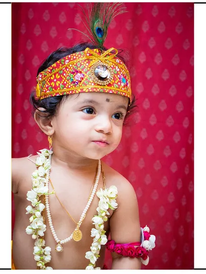 A beautiful profile shot from a Krishna photoshoot, highlighting the details of the traditional crown and jewelry against a rich red backdrop.