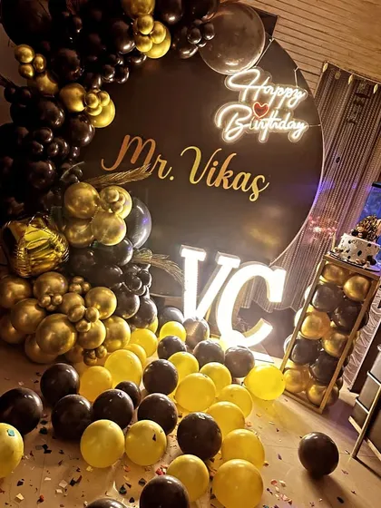 A sophisticated black and gold theme for 'Mr. Vikas'. This setup includes a custom circular backdrop, illuminated initials, and balloons scattered on the floor.