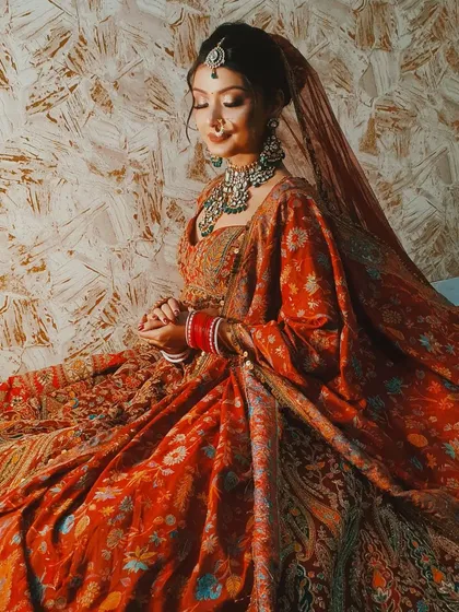 A candid, seated pose showing the rich fabric and embroidery of this maroon bridal lehenga. The green jewellery provides a beautiful contrast.