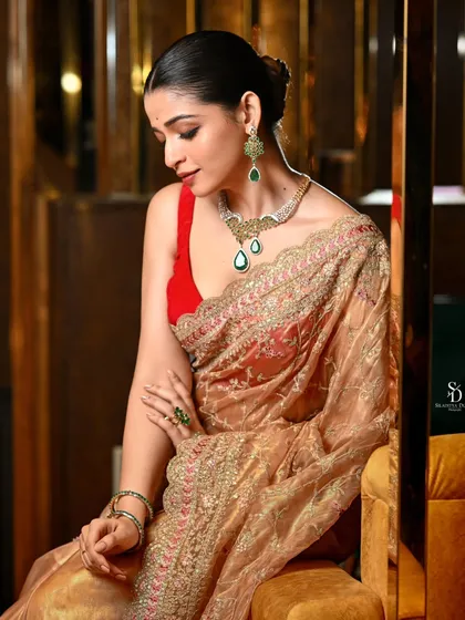 An elegant portrait of a model in a golden organza saree with emerald jewellery. Her demure, downward glance and the warm, luxurious lighting create a sophisticated and graceful mood.