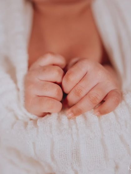A close-up of a newborn's tiny fists, nestled together. The soft texture of the white swaddle and the orange blanket create a cozy and warm feeling.
