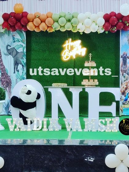 A wide view of a jungle-themed stage decoration. The setup includes a backdrop with animal prints, a neon 'Happy Birthday' sign, and large 'ONE' letters for a memorable first birthday.