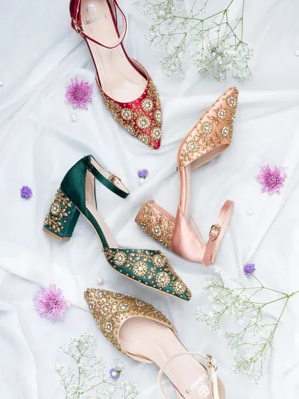 A beautiful flat lay of embellished ethnic footwear for women. The soft white fabric and scattered flowers create a dreamy and festive mood, perfect for a wedding collection.