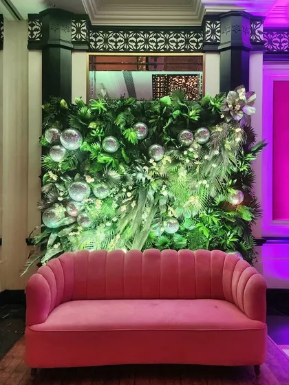 A chic photo spot combining a pink velvet sofa with a backdrop of lush greenery and silver baubles. The contrasting colors and textures create a modern and glamorous setting for guest photos.