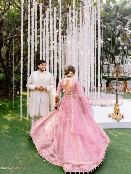The bride twirls in her beautiful pink lehenga as she approaches her groom, who waits for her amidst a curtain of white flowers. A moment of joyful anticipation.
