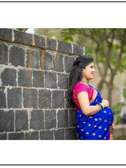 A contemplative side-profile of a mom-to-be in her beautiful blue saree, leaning against a stone wall.