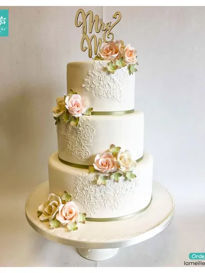 This classic three-tiered wedding cake exudes timeless elegance. It features intricate white-on-white lace piping, subtle ribbon bands, and is topped with a custom 'Mr & Mrs' topper and delicate sugar roses.