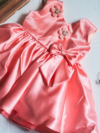 A beautiful salmon pink satin dress with a large bow and floral accents. This flat-lay shot highlights the rich color and luxurious texture of the fabric.