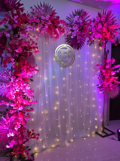 A vibrant pink floral arch against a white curtain backdrop with fairy lights. The golden "Tapasvi Amar Raho" sign adds a touch of elegance to this home ceremony setup.
