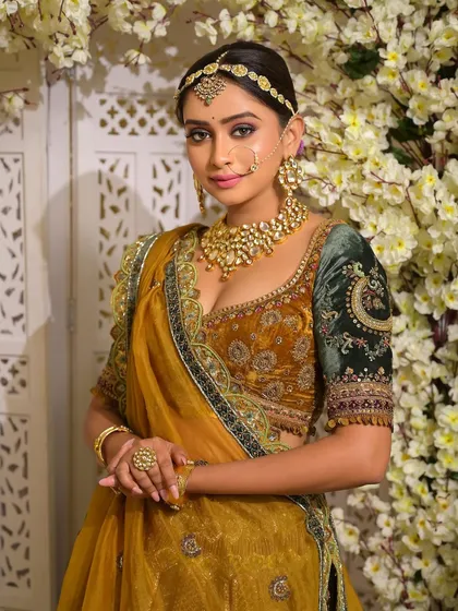 A classic bridal pose against a floral background. The makeup, with its soft smokey eye and flawless skin, complements the rich mustard and green tones of the traditional lehenga.