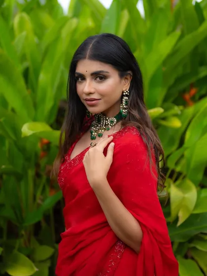 A close-up portrait of a model in a red saree, highlighting her stunning emerald necklace. The lush green background complements the jewelry and makes the entire image vibrant.