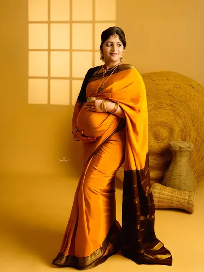 A simple and elegant studio portrait focusing on the beauty of a traditional saree. The warm yellow tones and soft window light create a serene and classic maternity photo.