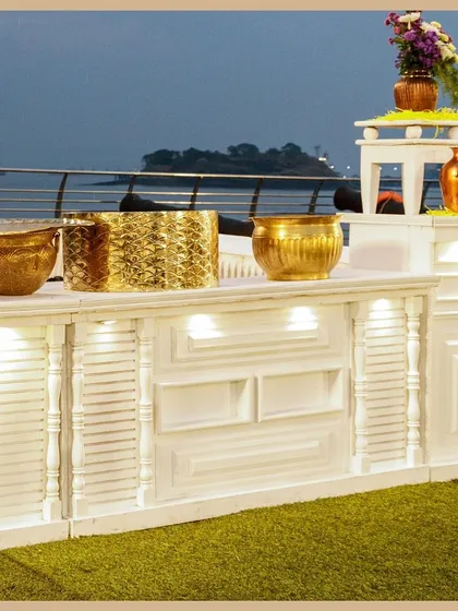 An elegant white buffet counter adorned with gleaming gold serving bowls and floral arrangements, set against a serene waterfront view. This is catering with a touch of class.