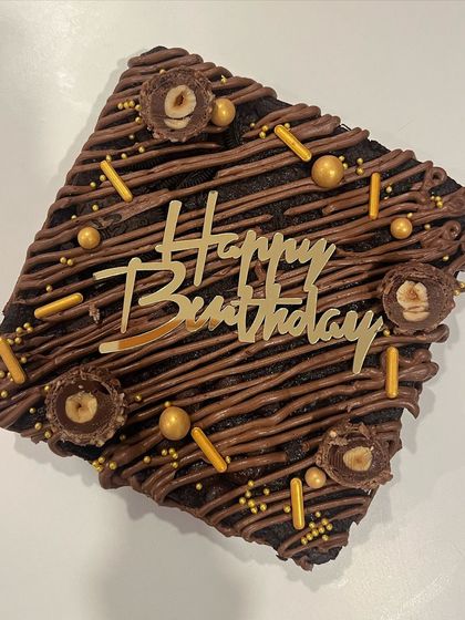 A close-up of my Ferrero Rocher brownie cake, perfect for a birthday celebration. It's drizzled with chocolate and topped with crunchy hazelnuts, sprinkles, and a "Happy Birthday" plaque.