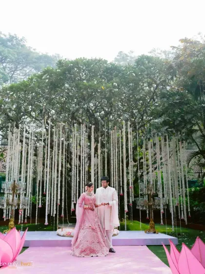 The couple stands together under a magnificent tree adorned with hanging floral strings, a grand and beautiful setting for their wedding ceremony.