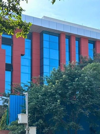 A closer shot of the red and blue glass facade, showing how the building interacts with its green surroundings. The design balances bold colors with natural elements.