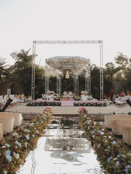 The wedding aisle at Ananta Udaipur, featuring a mirrored walkway lined with lush florals, leading to the elegant white mandap by the water.