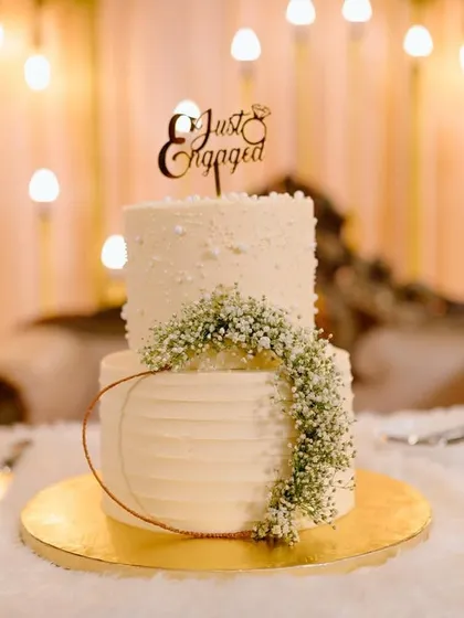 An elegant two-tiered cake for an engagement party, featuring a "Just Engaged" topper. The minimalist design is enhanced by a delicate cascade of baby's breath and a subtle textured buttercream finish.