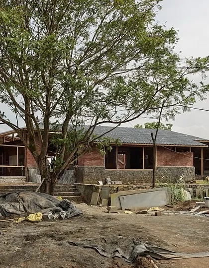 A wide view of the farmhouse under construction, nestled beside a large tree. The building's low-slung profile and use of natural materials help it settle into the rural Kamshet landscape.