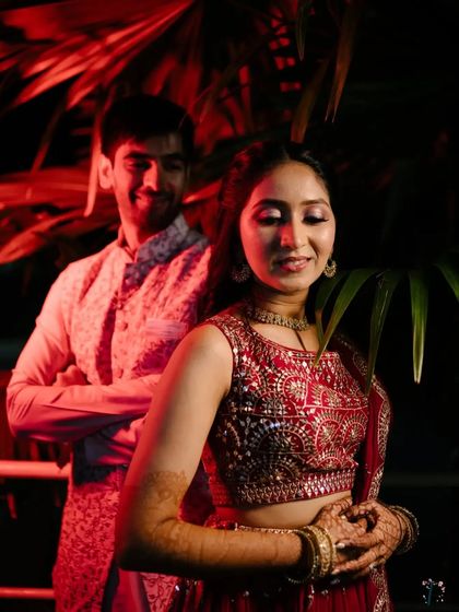 A stylish Sangeet portrait with dramatic red lighting, capturing the bride in the foreground with the groom smiling behind her.