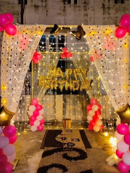 A terrace decoration with a fairy light curtain, "Happy Birthday" banner, and pink and white balloon columns.