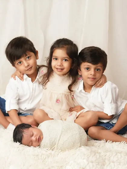 A beautiful sibling portrait where three older children in white and neutral tones are gathered lovingly around their new baby sibling, who is swaddled and sleeping peacefully on a soft white rug.