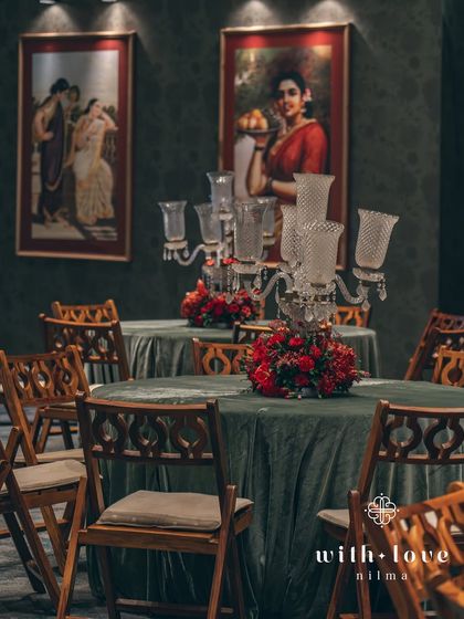 A dining table setup with a classic candelabra and red floral centerpiece. The entire scene is framed by the stunning Raja Ravi Varma paintings on the wall.