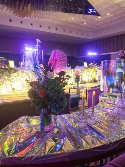 A lounge seating area with tables covered in shimmering, iridescent fabric. The decor is a mix of textures and finishes, from plush velvet sofas to reflective surfaces, all bathed in cool purple light.